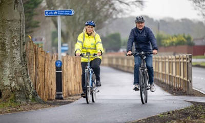 Councillor Joy Dennis rides along the new route on a bike and wearing a high visibility jacket. The MP Andrew Griffith rides alongside her on another bike, wearing a dark coloured jacket.