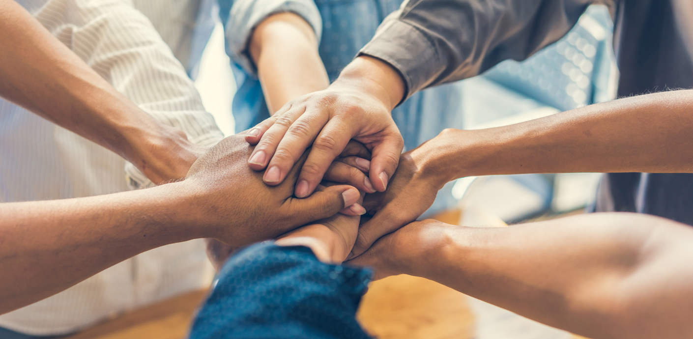 nojs A group of eight people in a circle each holding out one arm with their hands stacked on top of one another's in the middle of the circle.