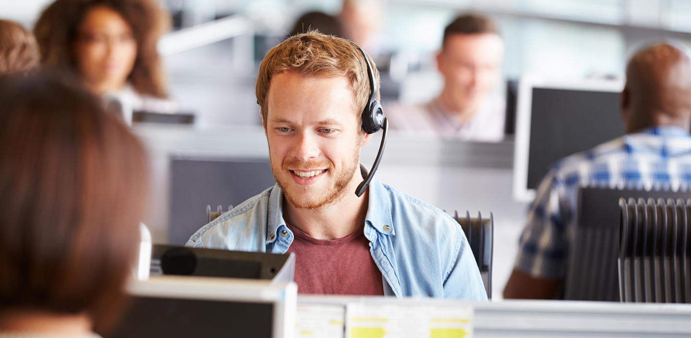 nojs A man wearing a telephony headset sat at a desk in a call centre.