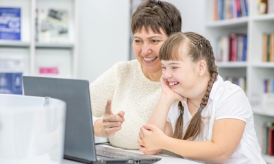 A lady sits with a young girl at a desk as they work on a laptop together.