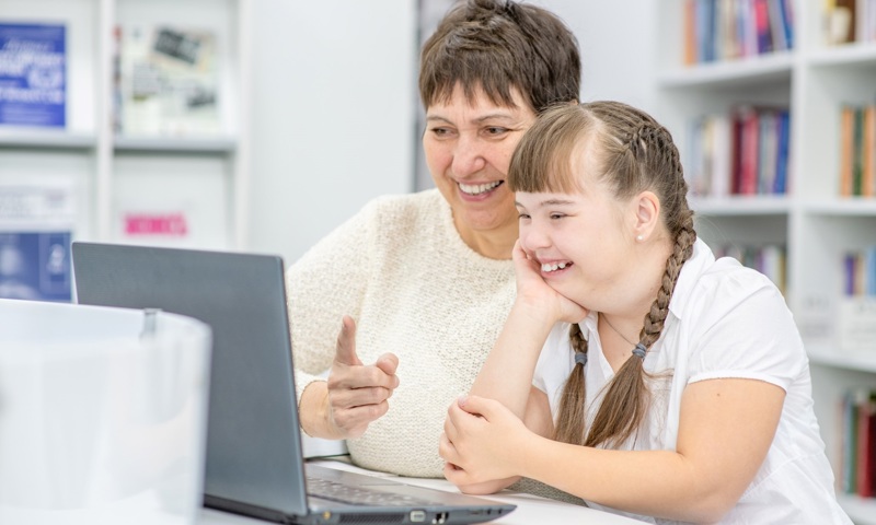 A lady sits with a young girl at a desk as they work on a laptop together.